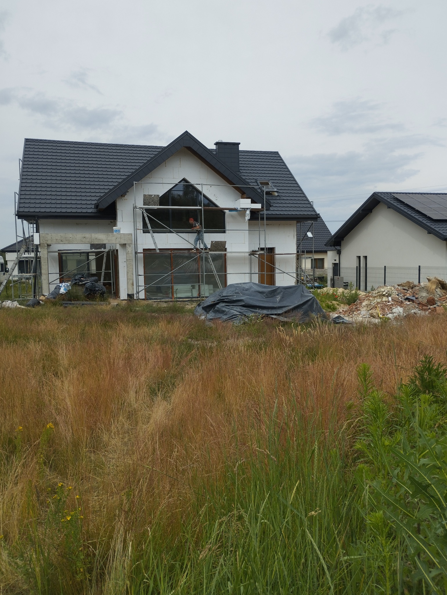 Dom w Kłobucku w trakcie ocieplania, widoczne rusztowania i pracownik na wysokości, w tle trawa i fragment sąsiedniego budynku.