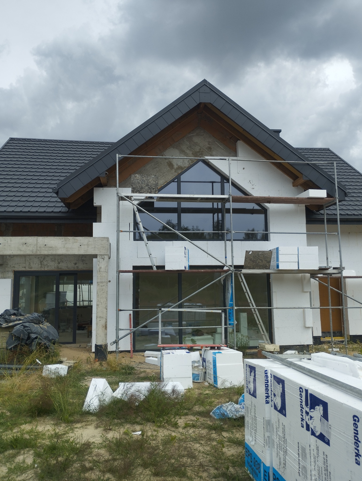 Dom jednorodzinny w trakcie ocieplania, widoczne rusztowania, materiały izolacyjne Genderka EPS 038 oraz częściowo ocieplona elewacja z charakterystycznym trójkątnym oknem na poddaszu.