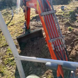 Widok z kabiny na pomarańczową koparkę Kubota z łyżką pełną ziemi w słoneczny dzień. Widoczne elementy sterujące i fragment ubrania operatora.