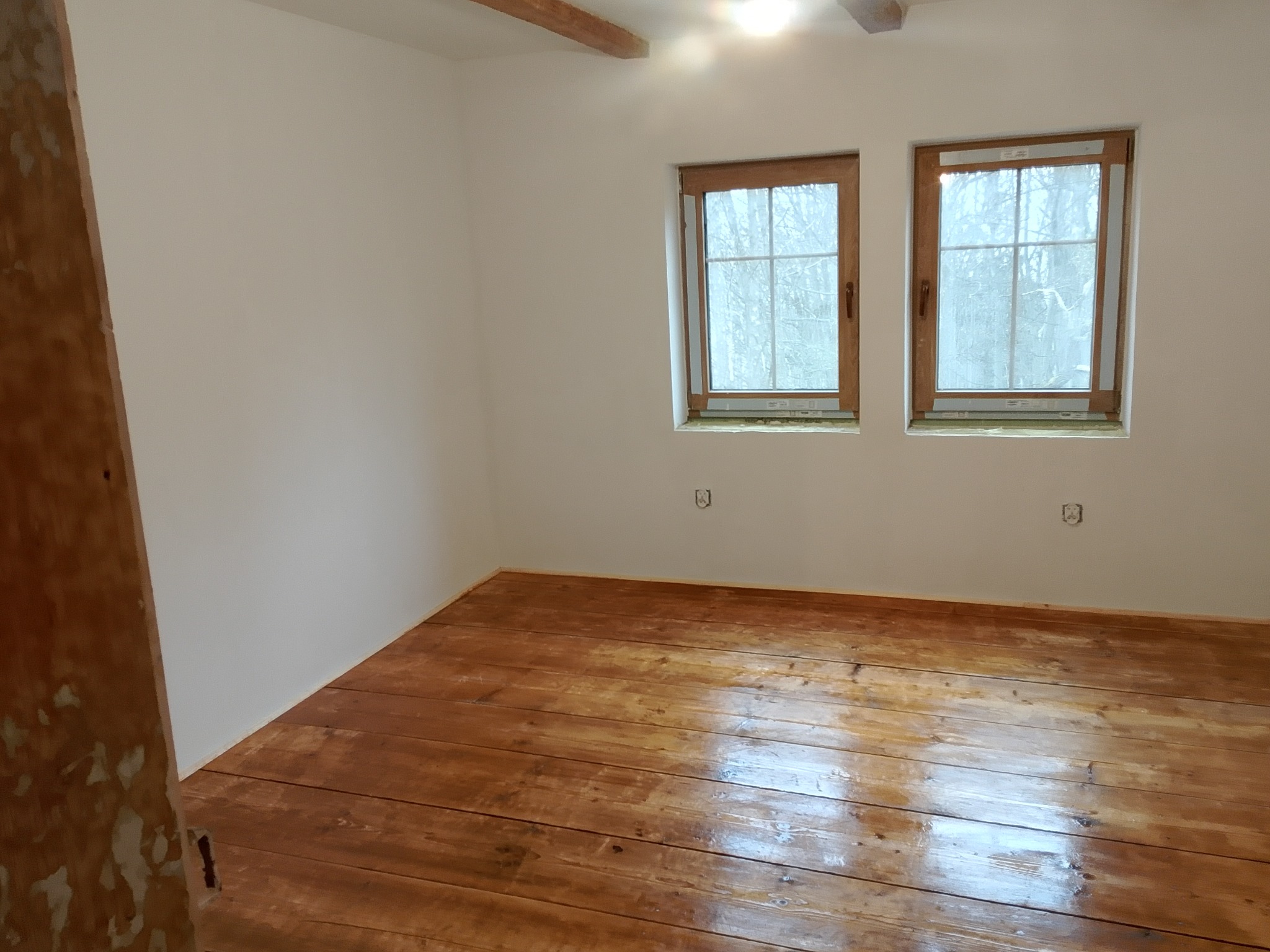 Wnętrze po renowacji: odnowiona drewniana podłoga z połyskiem, świeżo pomalowane białe ściany, dwa okna z drewnianymi ramami. Widoczny fragment drewnianej belki stropowej.