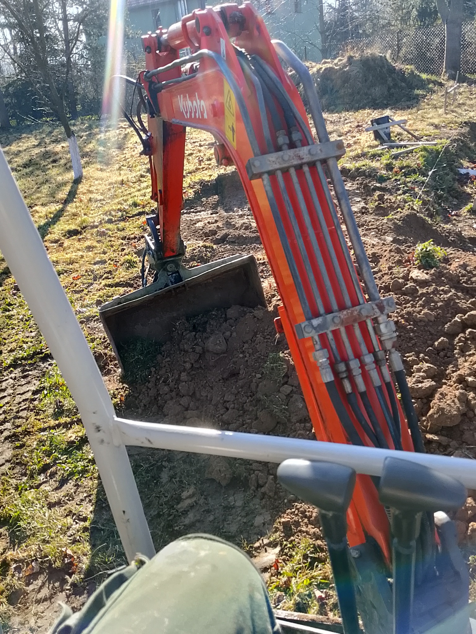 Widok z kabiny na pomarańczową koparkę Kubota z łyżką pełną ziemi w słoneczny dzień. Widoczne elementy sterujące i fragment ubrania operatora.