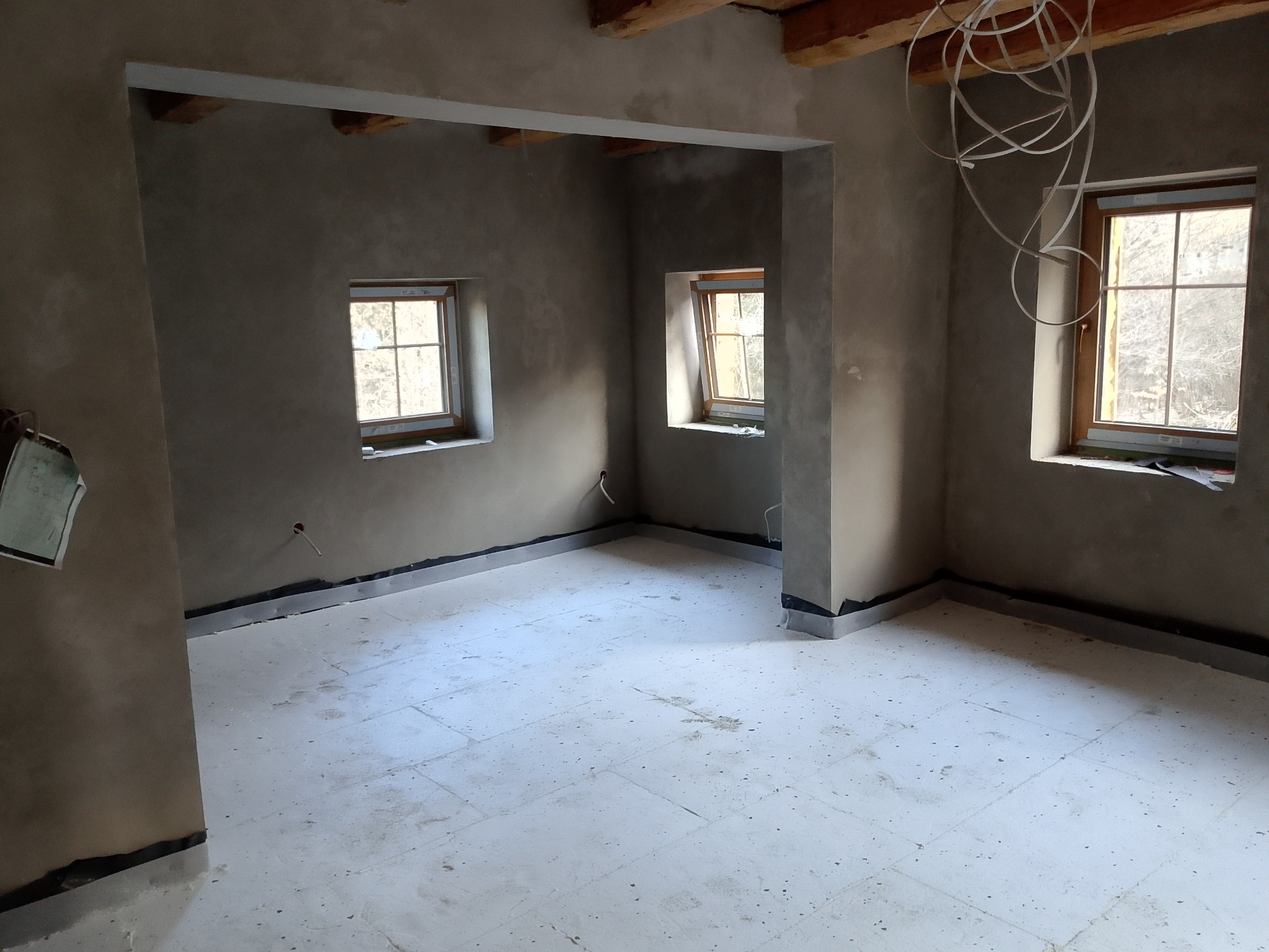 Surowy stan wnętrza: ściany po tynkowaniu, trzy okna z drewnianą ramą, widoczne kable pod sufitem, przygotowana podłoga. Wnętrze przed wykończeniem.