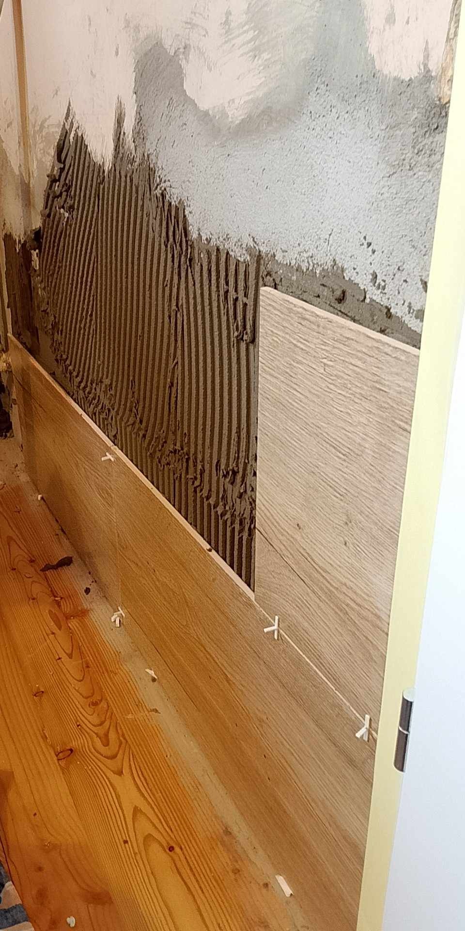 Układanie płytek imitujących drewno na ścianie. Widoczna warstwa kleju, płytka i krzyżyki dystansowe. Parkiet zabezpieczony przed zabrudzeniem.