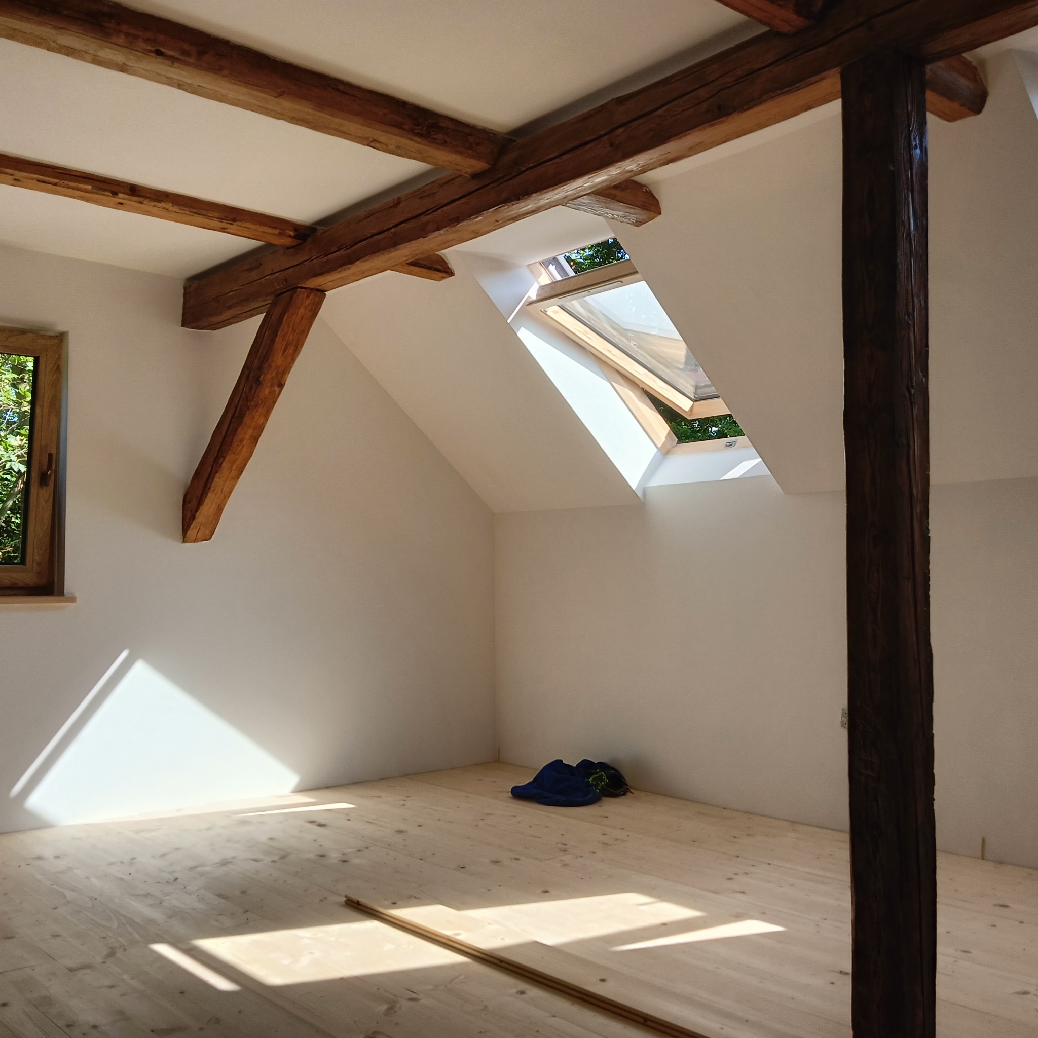 Jasne poddasze po remoncie z drewnianymi belkami, oknem dachowym i podłogą. Na podłodze leżą niebieskie ubrania. Widoczne promienie słońca.