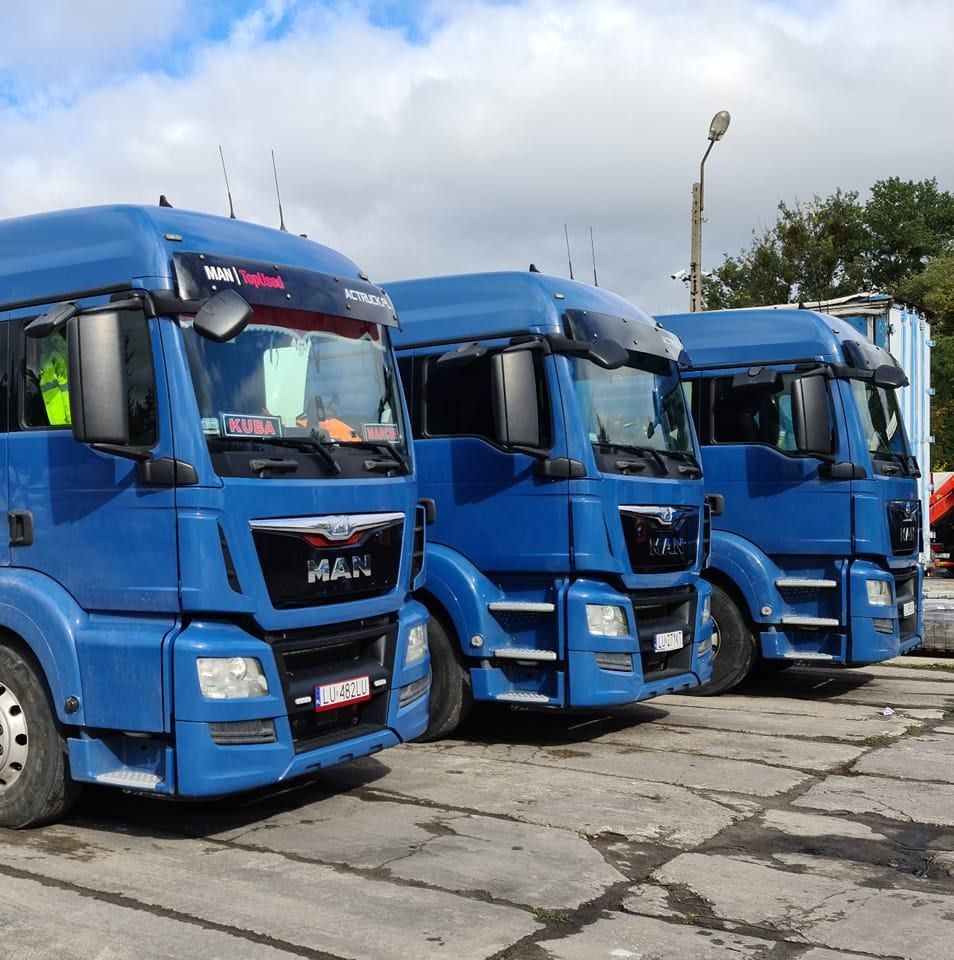 Trzy niebieskie ciężarówki MAN zaparkowane obok siebie na betonowym placu, widoczne tablice rejestracyjne i nazwy kierowców na przedniej szybie.