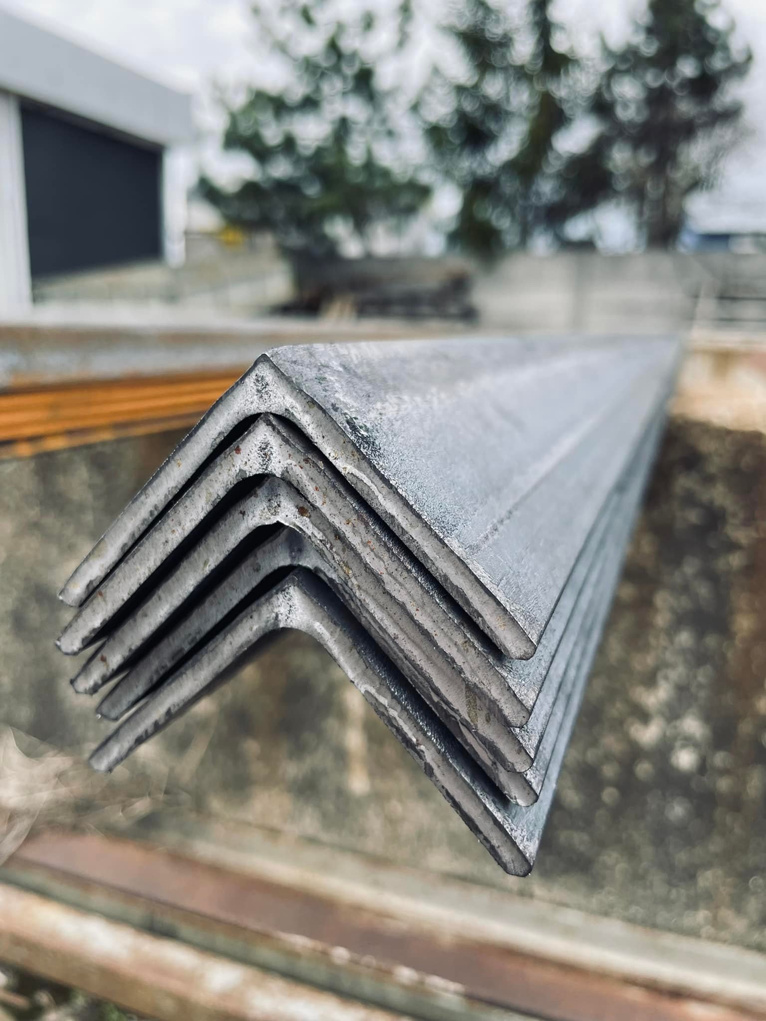 Stos kątowników stalowych ułożonych jeden na drugim, widoczne ślady korozji, tło rozmyte z budynkiem i drzewem.