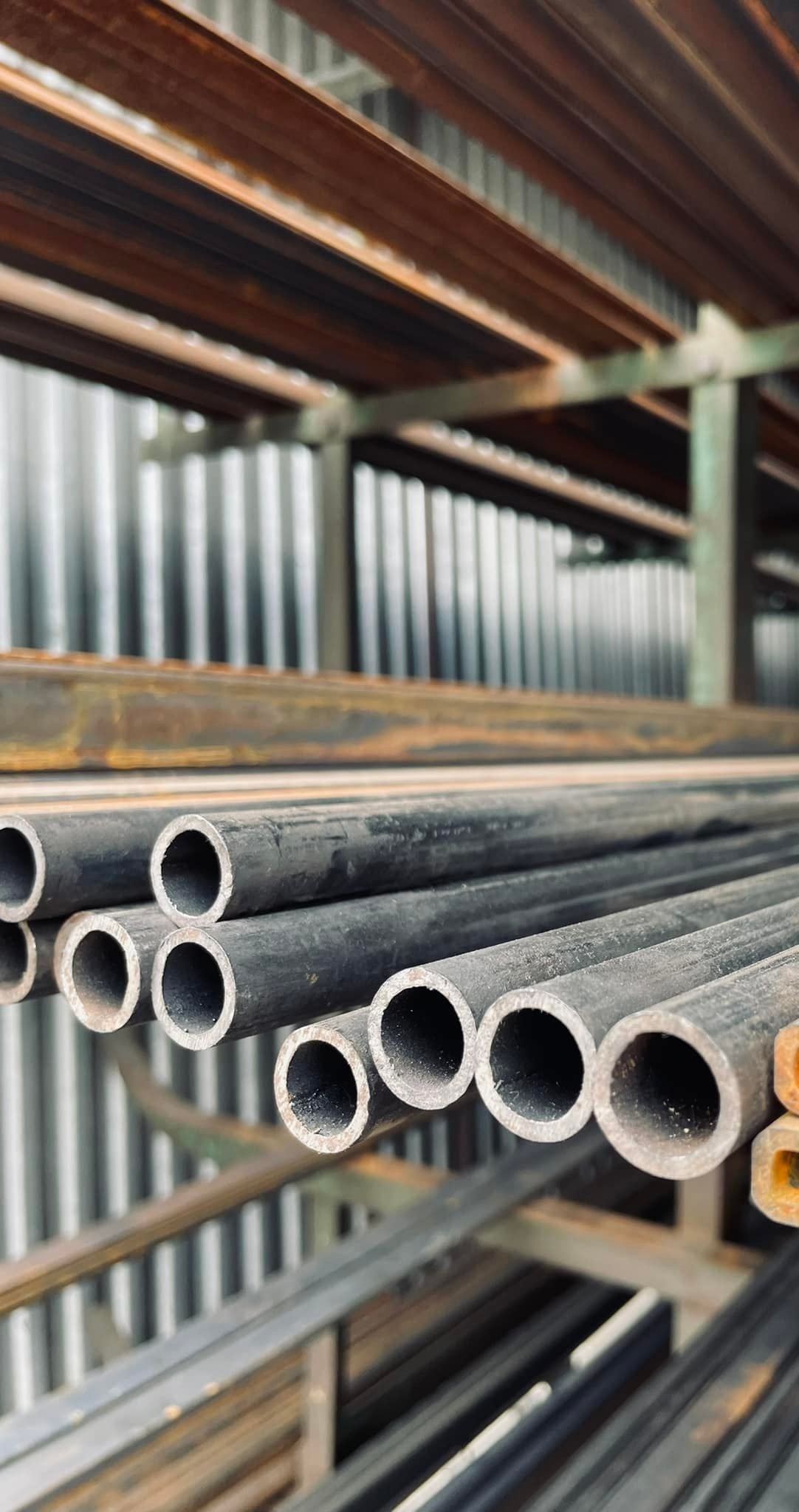 Stos rur stalowych o okrągłym przekroju na metalowym regale, widoczne ślady rdzy na niektórych elementach konstrukcyjnych.