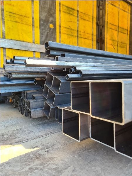 Stalowe profile kwadratowe ułożone w stos, widoczne żółte drzwi kontenera w tle.