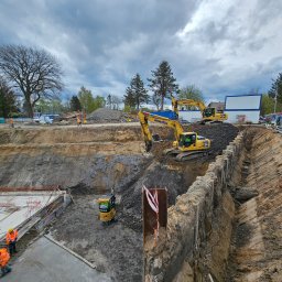 Patryk Janicki Usługi Budowlane - Szeroki wykop pod budowę z trzema koparkami Komatsu, widoczne zbrojenia fundamentów i pracownicy w pomarańczowych kamizelkach, pochmurne niebo w tle.