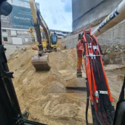 Widok z kabiny na żółtą koparkę pracującą na placu budowy, z widocznym wykopem i stertą piasku. W tle budynek z reklamą.