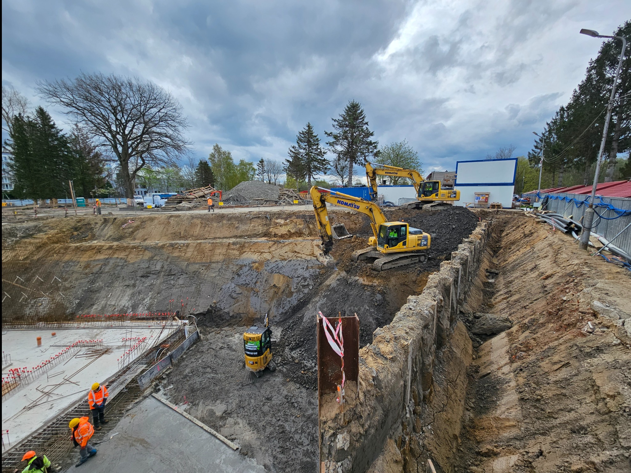 Szeroki wykop pod budowę z trzema koparkami Komatsu, widoczne zbrojenia fundamentów i pracownicy w pomarańczowych kamizelkach, pochmurne niebo w tle.