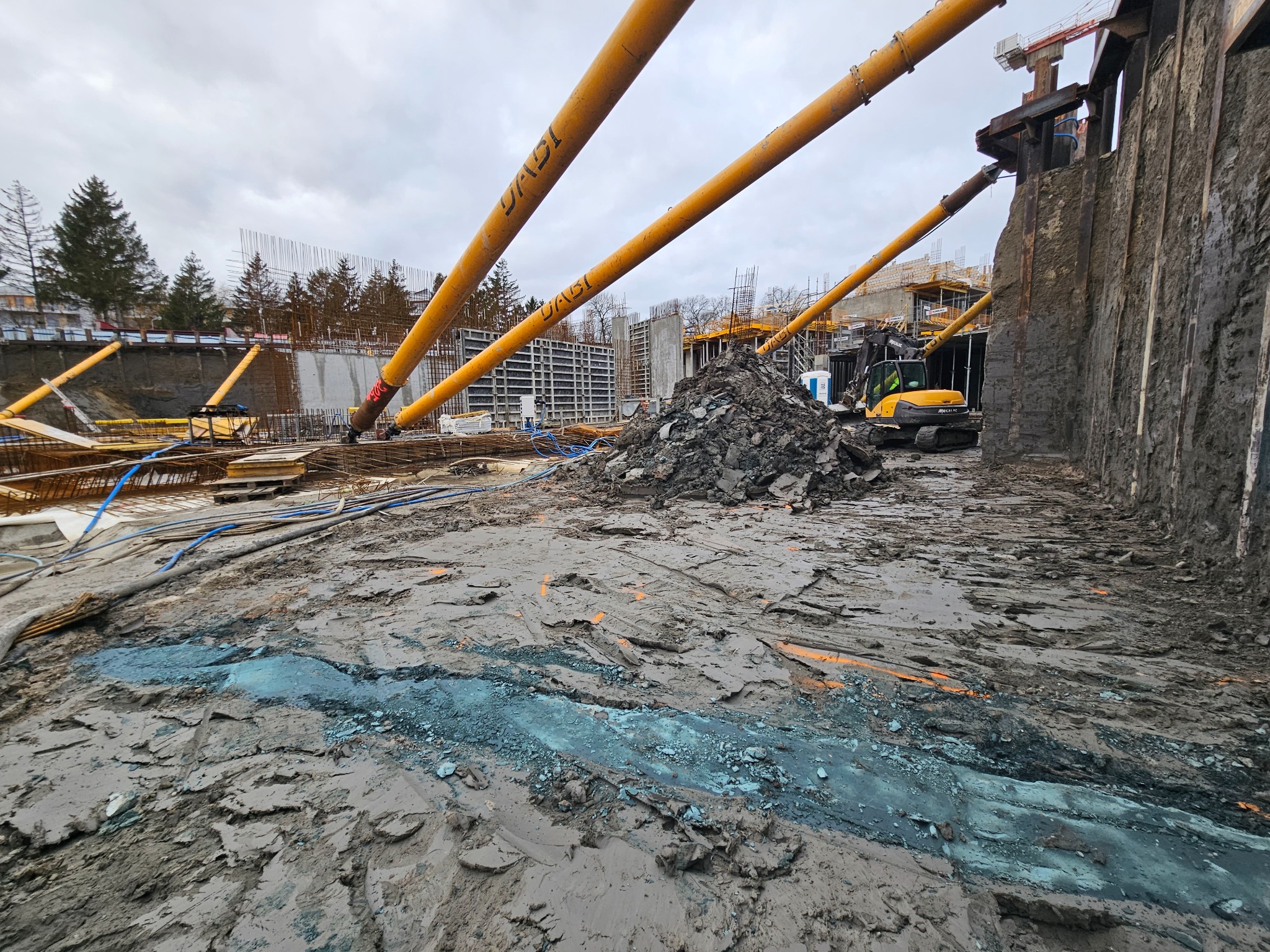 Szeroki kadr na plac budowy z widocznym wykopem zabezpieczonym ścianką berlińską, stertą ziemi i pracującą koparką; żółte rury wzmacniające konstrukcję nachylone pod kątem.