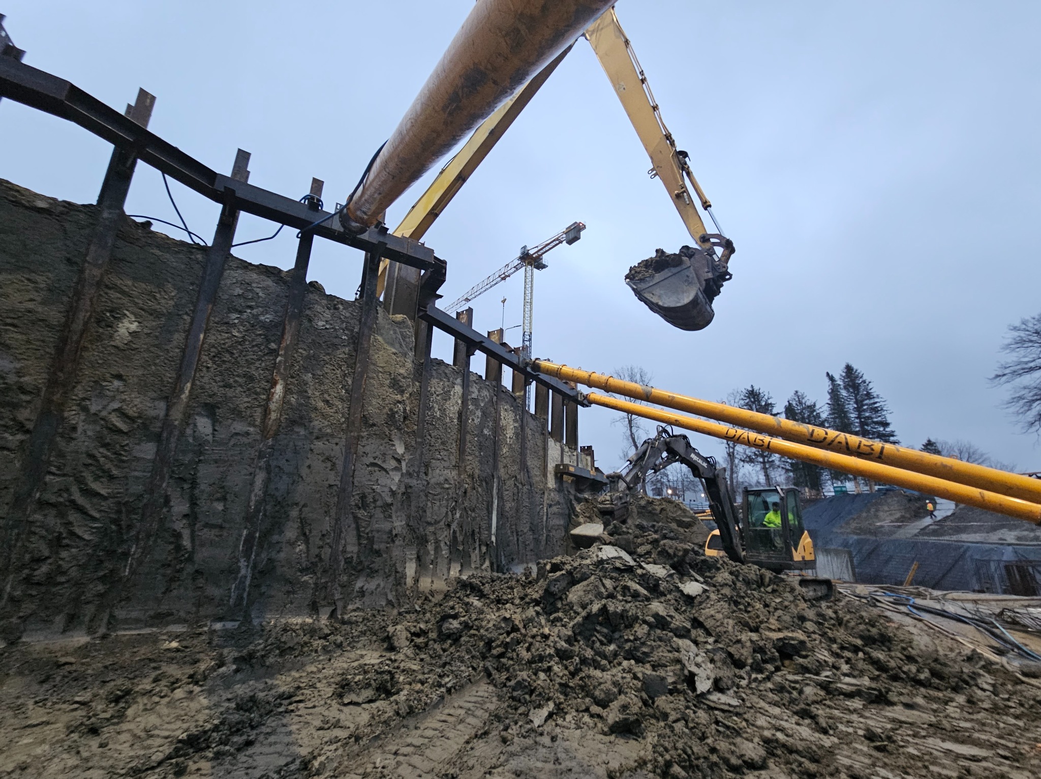 Głęboki wykop zabezpieczony stalowymi panelami, z pracującą koparką i widocznym dźwigiem w tle.