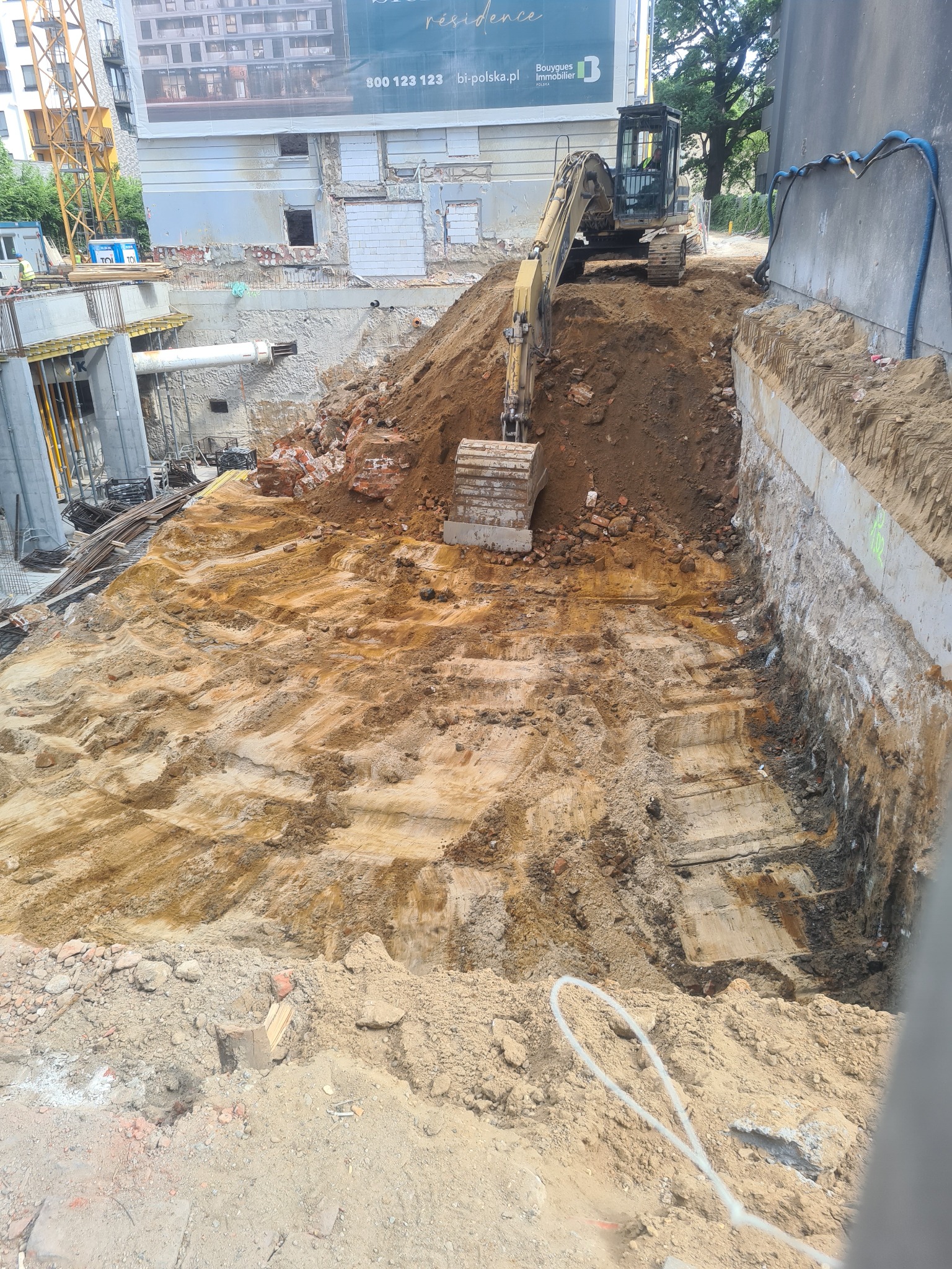 Żółta koparka na placu budowy, widok z góry na wykop z fragmentami cegieł i gruzu, w tle budynek z reklamą i dźwig budowlany.