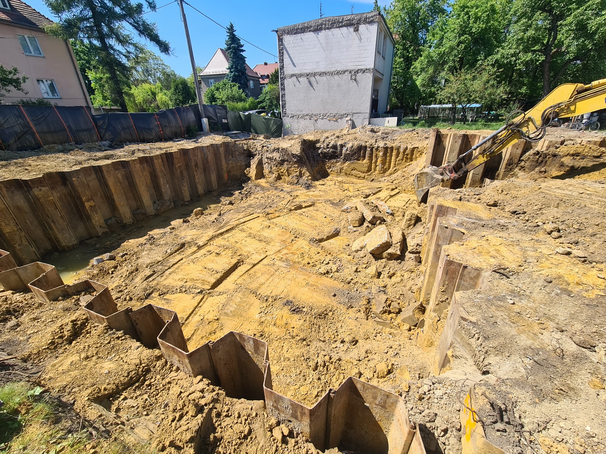 Głęboki wykop zabezpieczony ściankami Larsena, z żółtą koparką w tle, częściowo rozebrany budynek w tle, słoneczny dzień.
