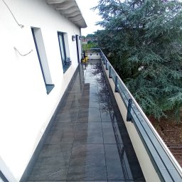 Damian Dawid - Wyłożony ciemnymi płytkami taras z szarą balustradą, widok na duży iglasty krzew.