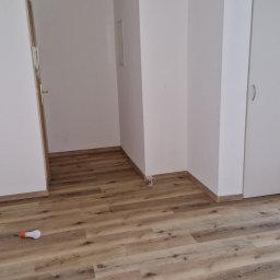 Damian Dawid - Jasne panele podłogowe w nowym pomieszczeniu, widoczna żarówka na podłodze i kubek przy ścianie, świeżo po ułożeniu.