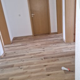 Damian Dawid - Nowo położone panele podłogowe imitujące drewno w jasnym odcieniu, widoczne drzwi w tle i biała ściana, na podłodze leży żarówka halogenowa.