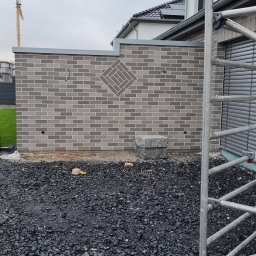 Damian Dawid - Świeżo wymurowana ściana z szarej cegły klinkierowej z dekoracyjnym wzorem rombu, widoczne przygotowane podłoże z czarnego grysu i fragment rusztowania.