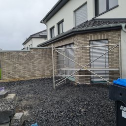 Damian Dawid - Szara kostka elewacyjna na ścianie domu z dekoracyjnym wzorem w kształcie rombu, widoczne rusztowanie i czarne kamienie na podłożu.