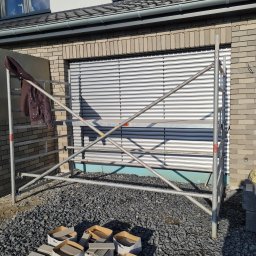 Damian Dawid - Metalowe rusztowanie przed elewacją budynku z szarej cegły i zamkniętą żaluzją okienną, na rusztowaniu brudna kurtka robocza, na ziemi gruz i kartony z płytkami elewacyjnymi.