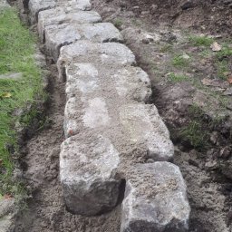 Damian Dawid - Rząd ułożonych, szarych kostek brukowych oddzielających trawnik od ziemi, z dwoma kostkami leżącymi obok, zasypane piaskiem.