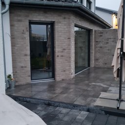 Damian Dawid - Nowoczesny taras z jasnej cegły z dużymi, przesuwnymi oknami i wyłożony szarymi płytami, częściowo pokryty kostką brukową w odcieniach szarości, widok na dom jednorodzinny w pochmurny dzień.