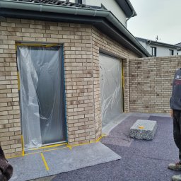 Damian Dawid - Przygotowania do malowania elewacji domu z cegły: okna i drzwi zabezpieczone folią i taśmą malarską, podłoże wyłożone matą ochronną.