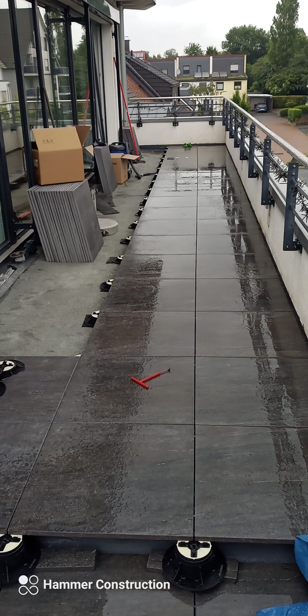 Montaż płytek tarasowych na regulowanych wspornikach, widoczne narzędzie do wyrównywania fug, częściowo ułożona powierzchnia, tło stanowią budynki mieszkalne.