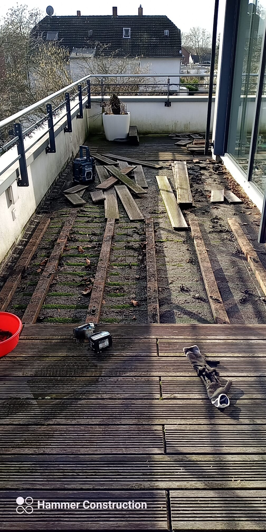 Demontaż drewnianego tarasu na balkonie, widoczne legary, częściowo usunięte deski, narzędzia akumulatorowe i rękawice robocze. W tle budynek mieszkalny.