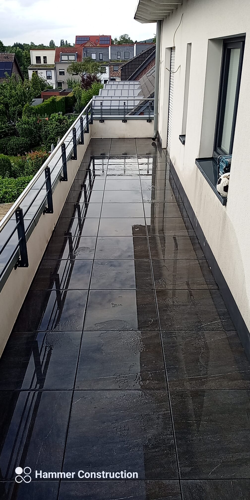 Długi balkon z ciemnymi, błyszczącymi płytkami, odbijającymi niebo, z czarną balustradą i piłką do nogi wystającą z okna.