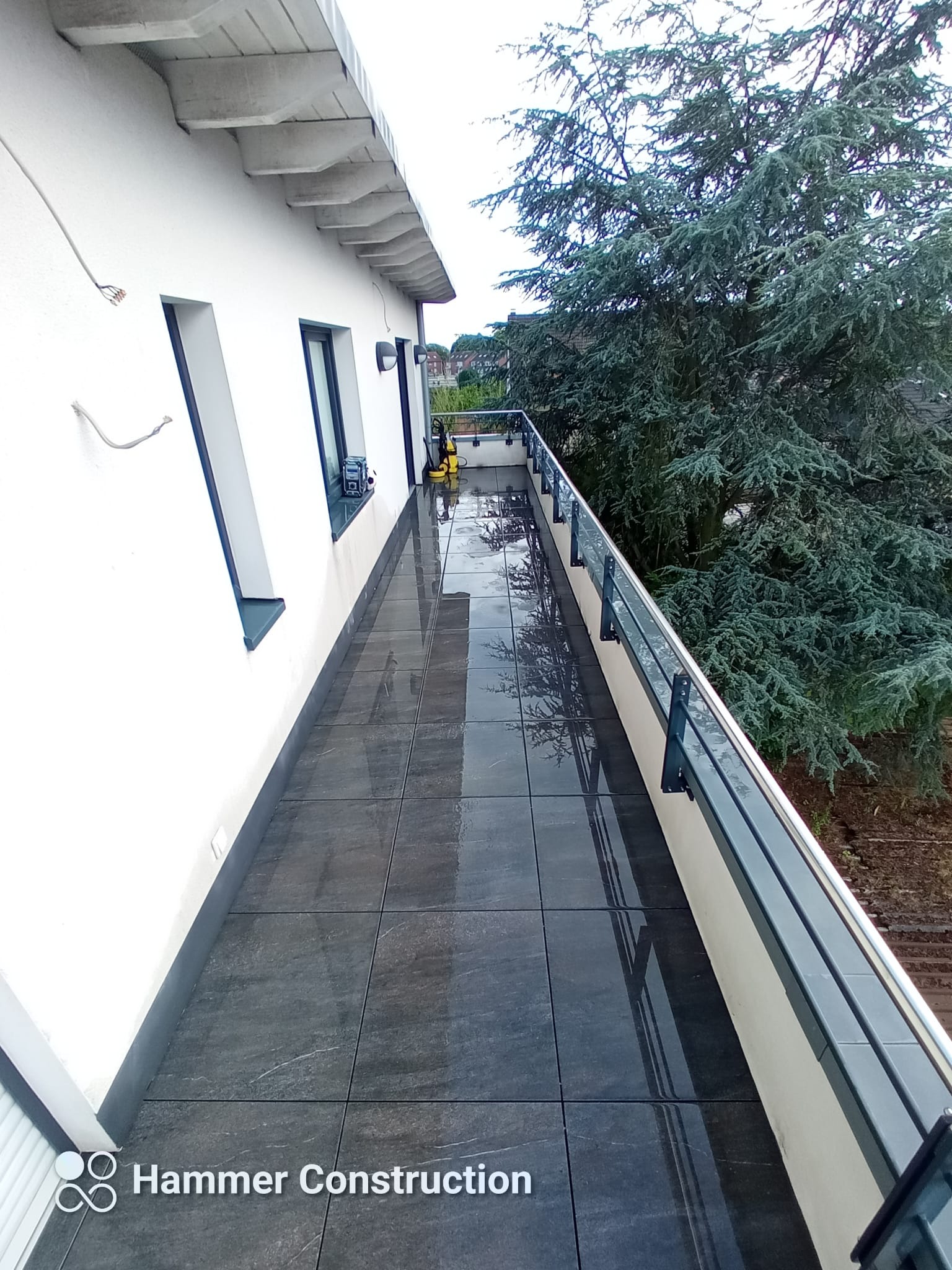 Wyłożony ciemnymi płytkami taras z szarą balustradą, widok na duży iglasty krzew.