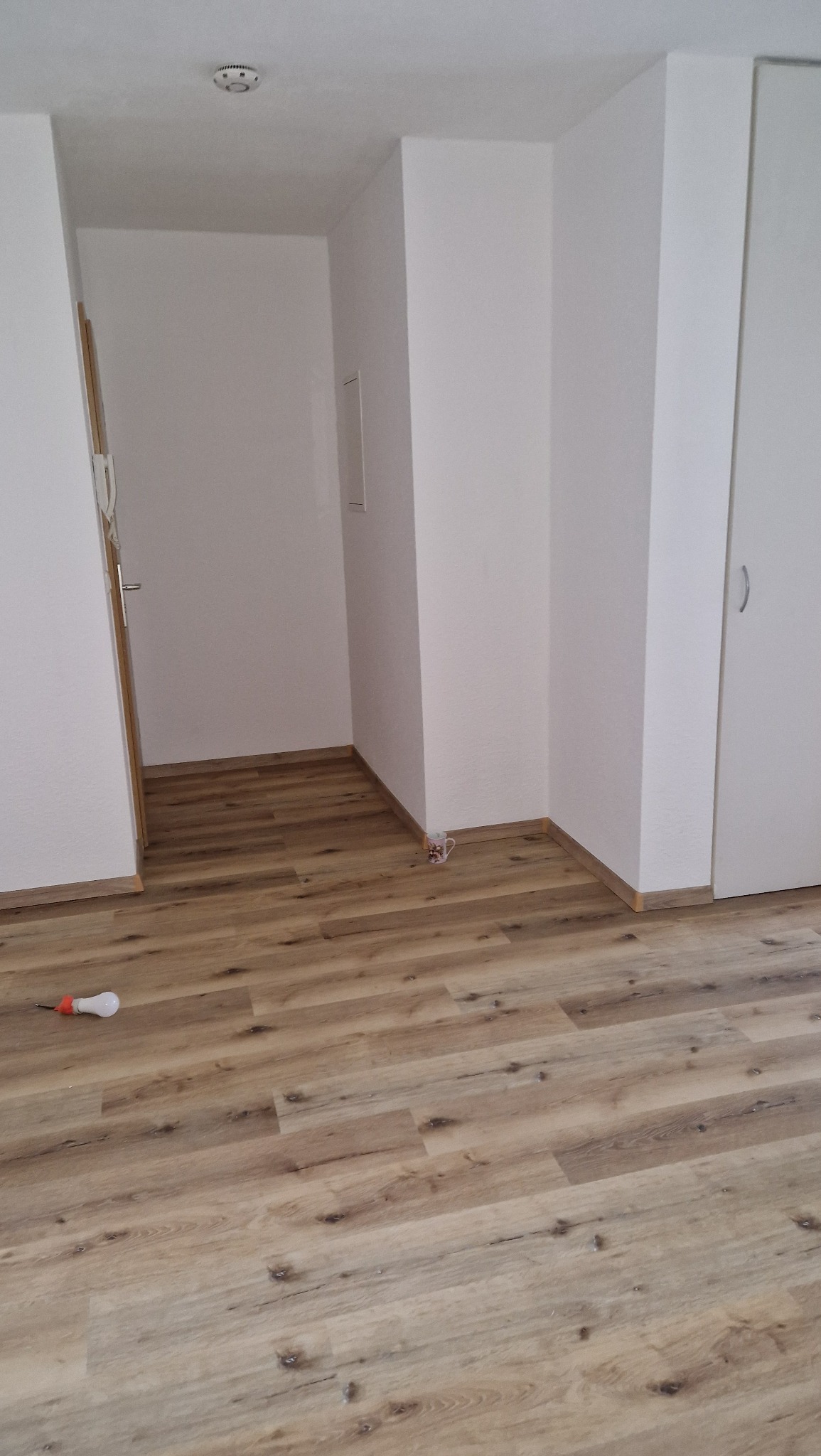 Jasne panele podłogowe w nowym pomieszczeniu, widoczna żarówka na podłodze i kubek przy ścianie, świeżo po ułożeniu.