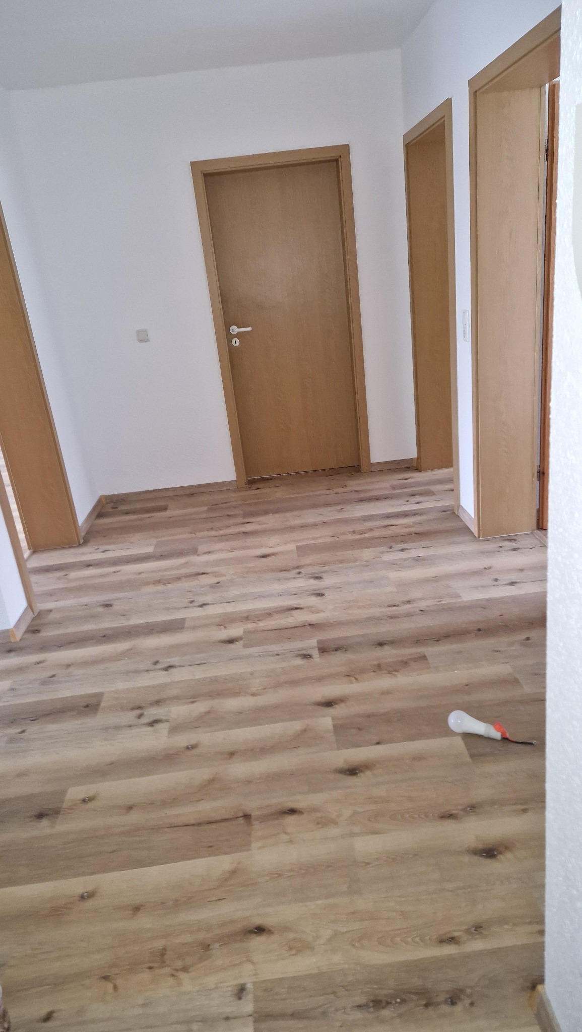 Nowo położone panele podłogowe imitujące drewno w jasnym odcieniu, widoczne drzwi w tle i biała ściana, na podłodze leży żarówka halogenowa.