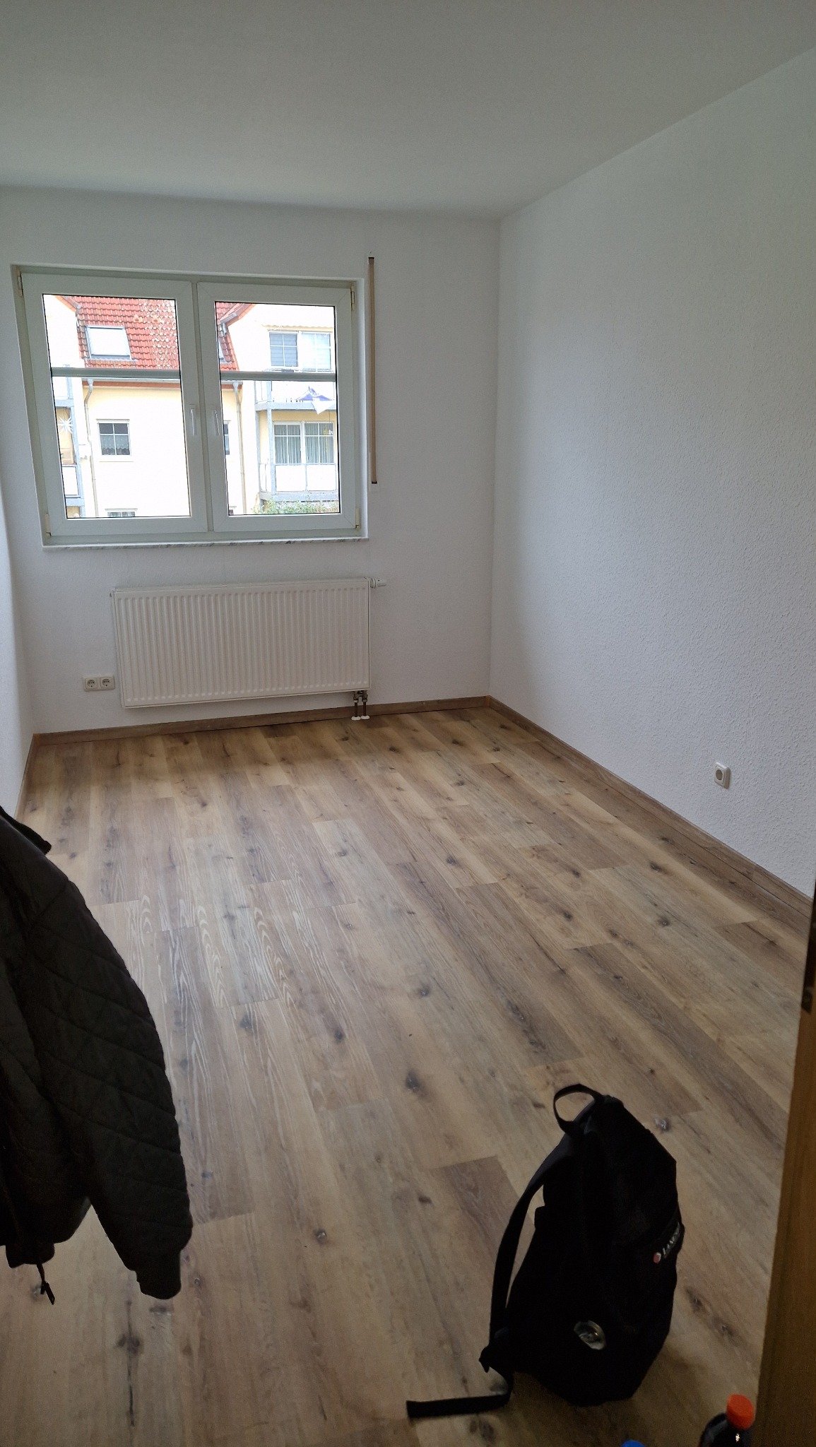 Pusty pokój po remoncie z nową podłogą z paneli, widoczny grzejnik pod oknem i czarny plecak na podłodze.