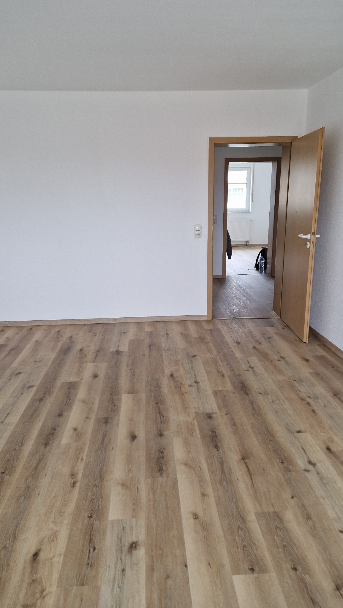 Jasnobrązowe panele podłogowe ułożone w pustym pokoju z białymi ścianami i otwartymi drewnianymi drzwiami do sąsiedniego pomieszczenia.