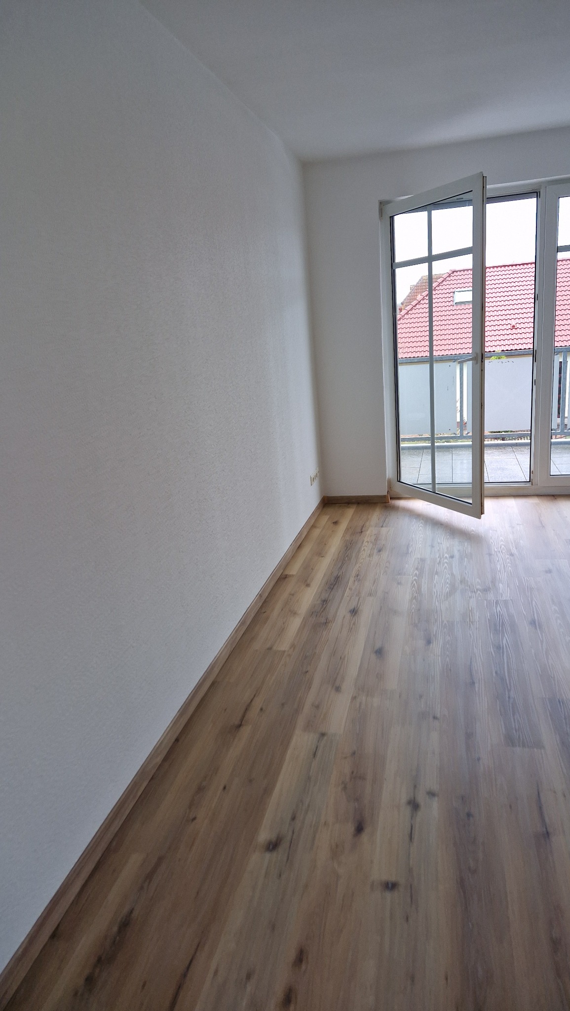 Wnętrze pustego pokoju z nową, jasną podłogą z paneli, białe ściany i otwarte drzwi balkonowe z widokiem na czerwony dach budynku.