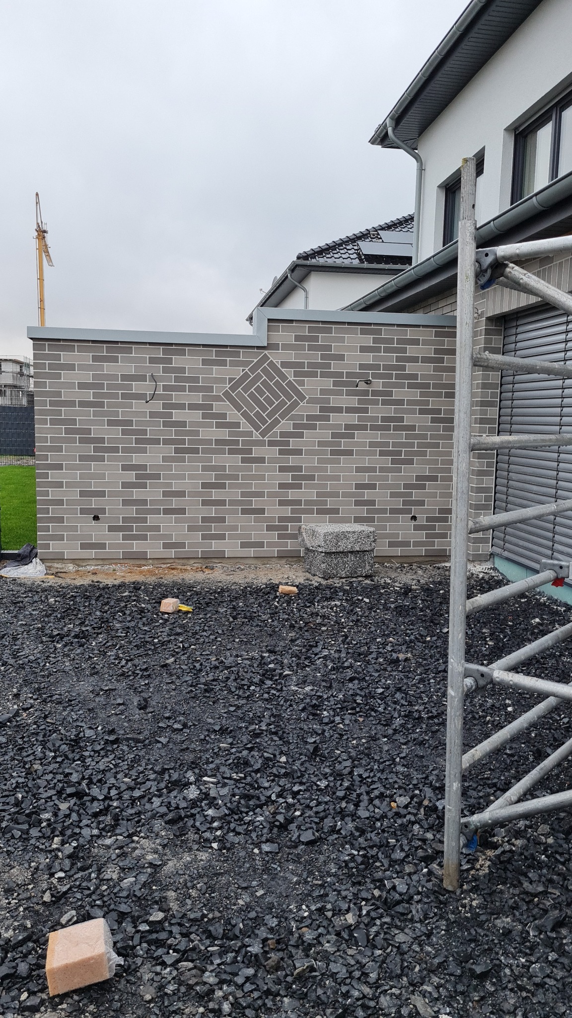 Świeżo wymurowana ściana z szarej cegły klinkierowej z dekoracyjnym wzorem rombu, widoczne przygotowane podłoże z czarnego grysu i fragment rusztowania.