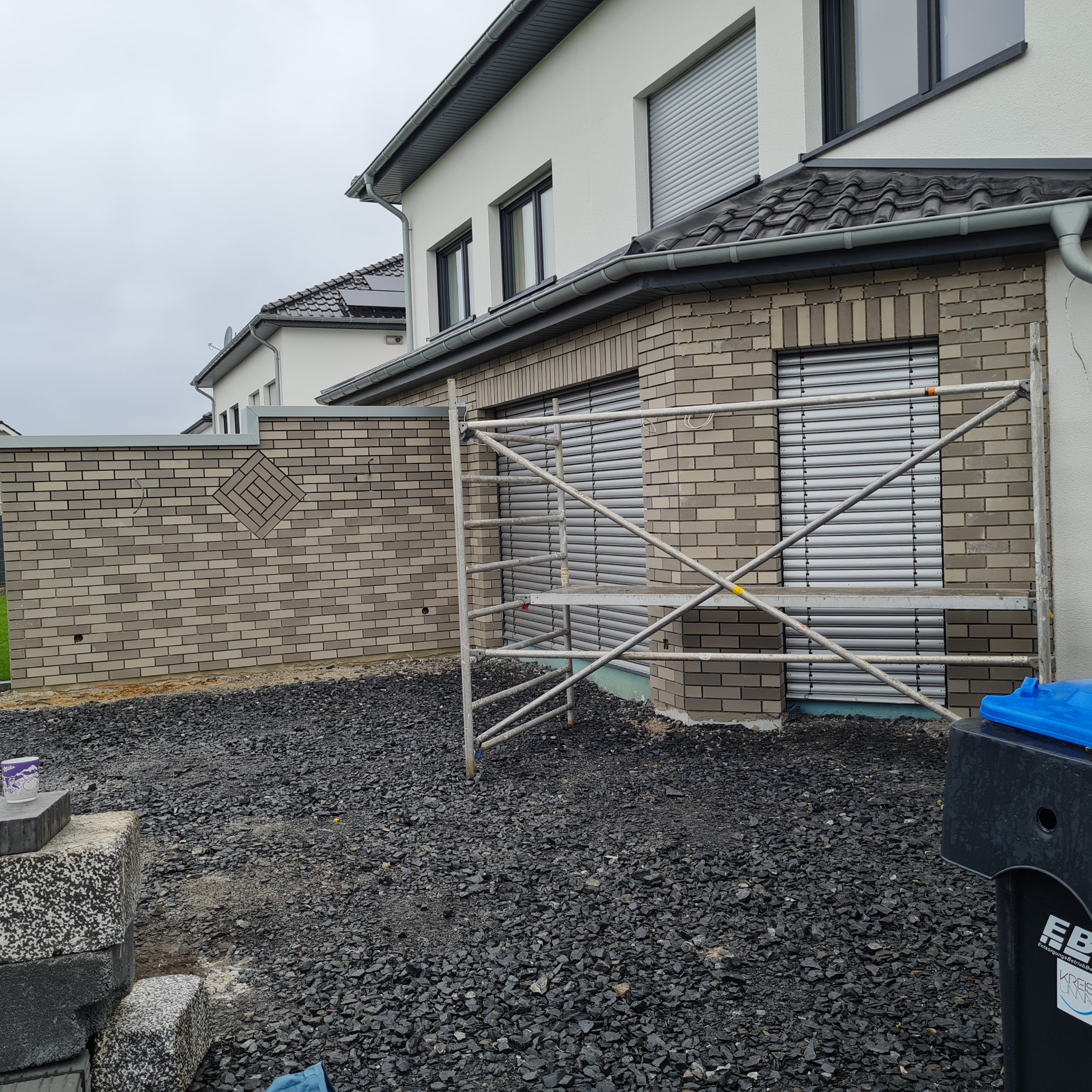 Szara kostka elewacyjna na ścianie domu z dekoracyjnym wzorem w kształcie rombu, widoczne rusztowanie i czarne kamienie na podłożu.