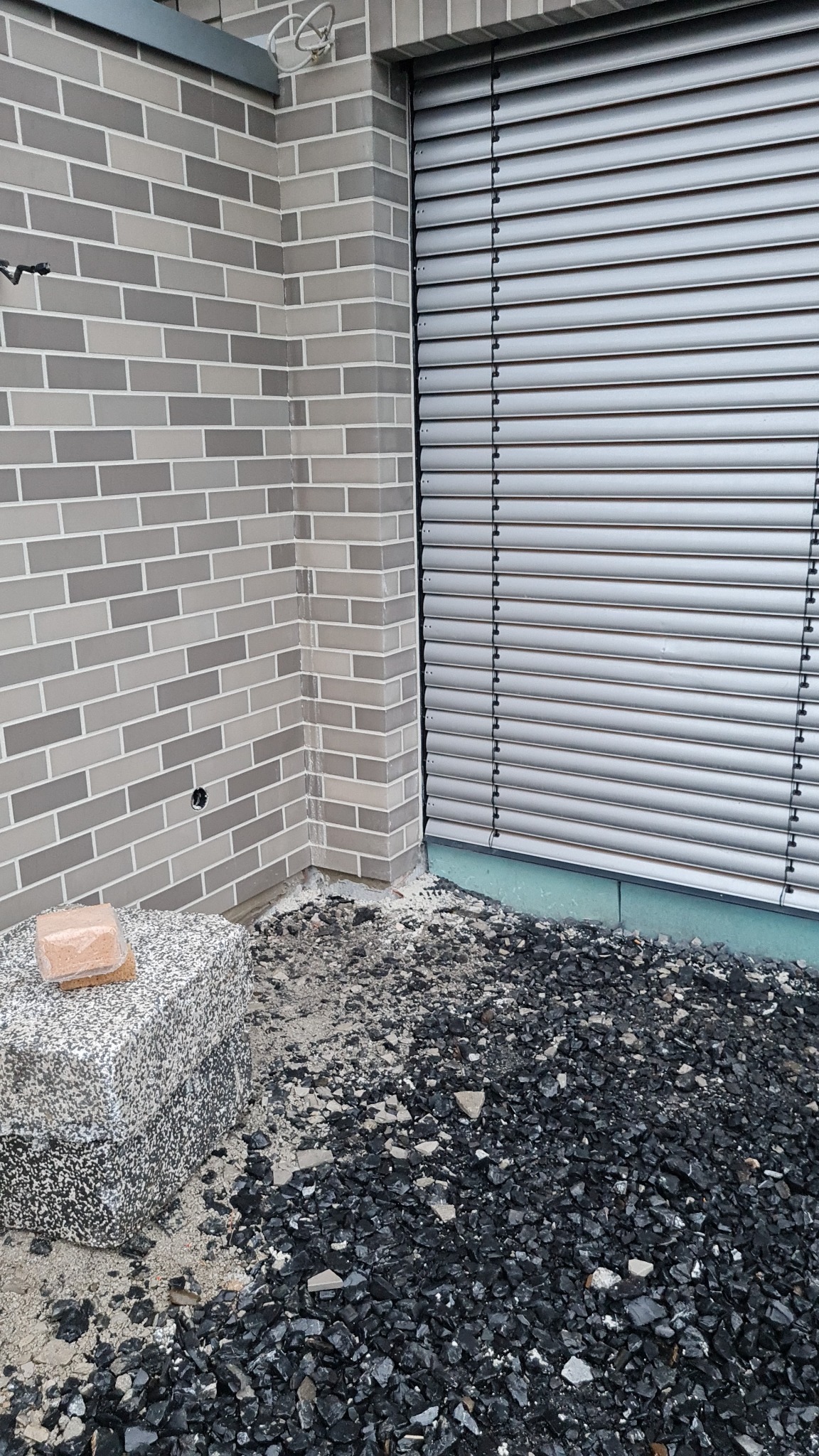 Szara ściana z cegły klinkierowej, zasłonięte żaluzje okienne, kostka granitowa i wysypany tłuczeń kamienny u podstawy ściany.