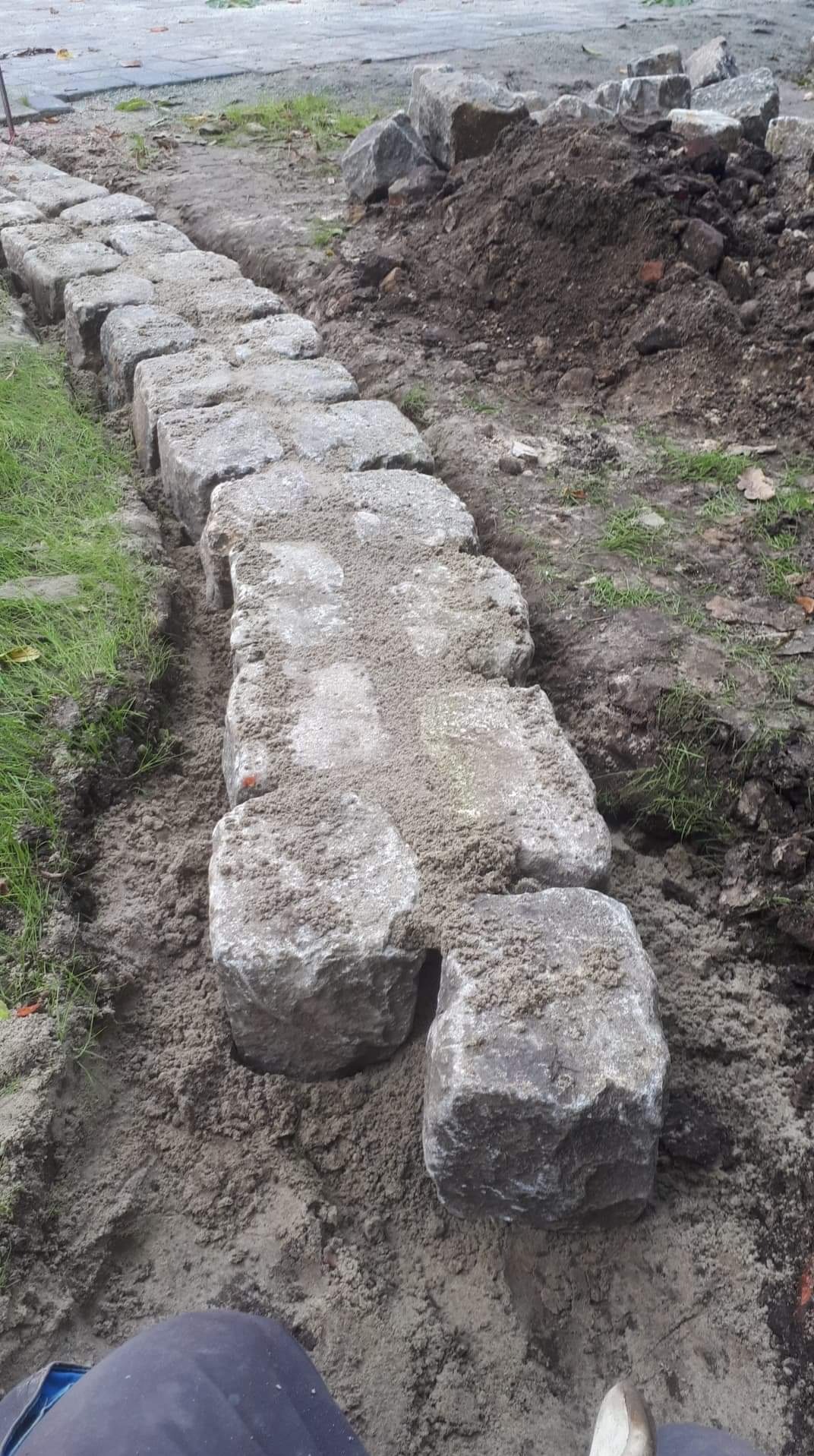 Rząd ułożonych, szarych kostek brukowych oddzielających trawnik od ziemi, z dwoma kostkami leżącymi obok, zasypane piaskiem.