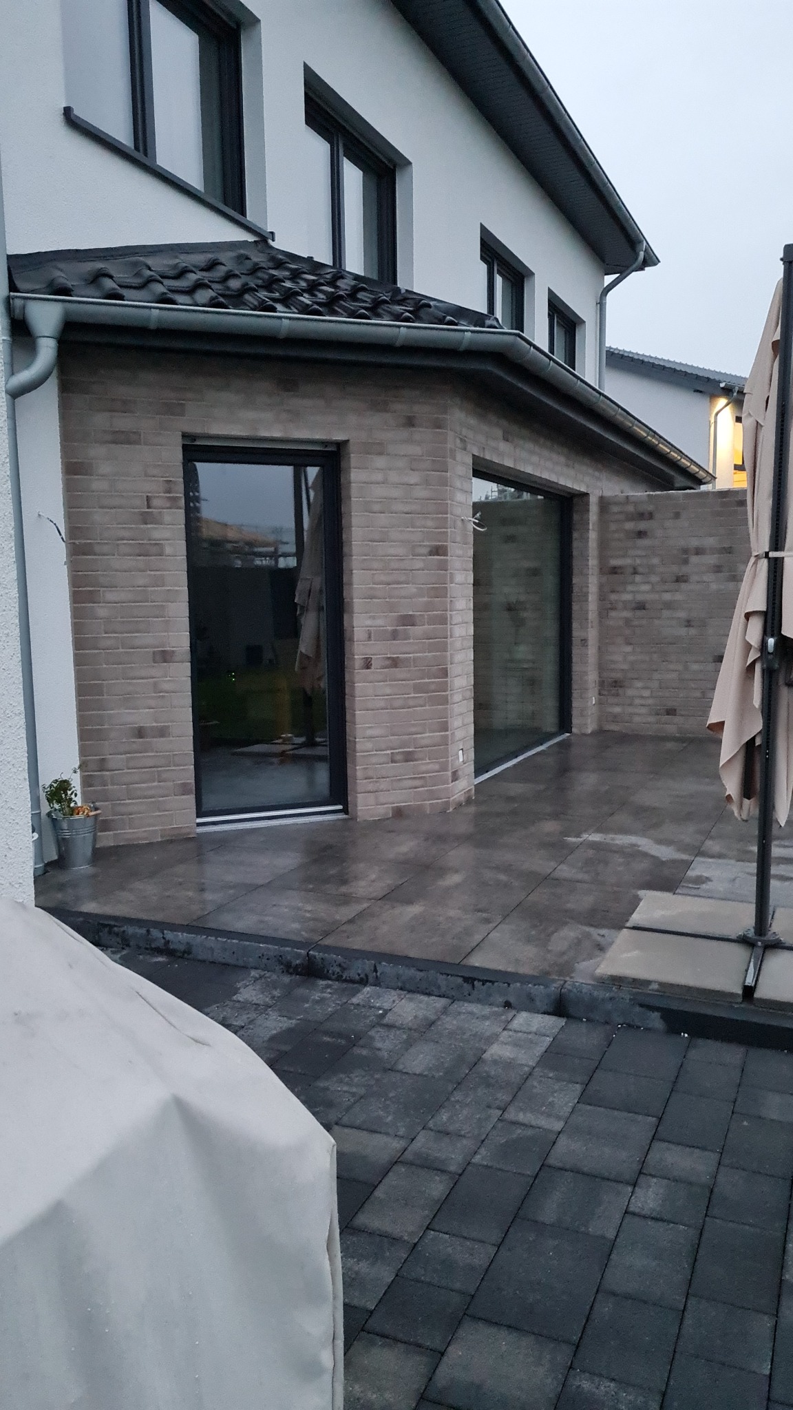 Nowoczesny taras z jasnej cegły z dużymi, przesuwnymi oknami i wyłożony szarymi płytami, częściowo pokryty kostką brukową w odcieniach szarości, widok na dom jednorodzinny w pochmurny dzień.