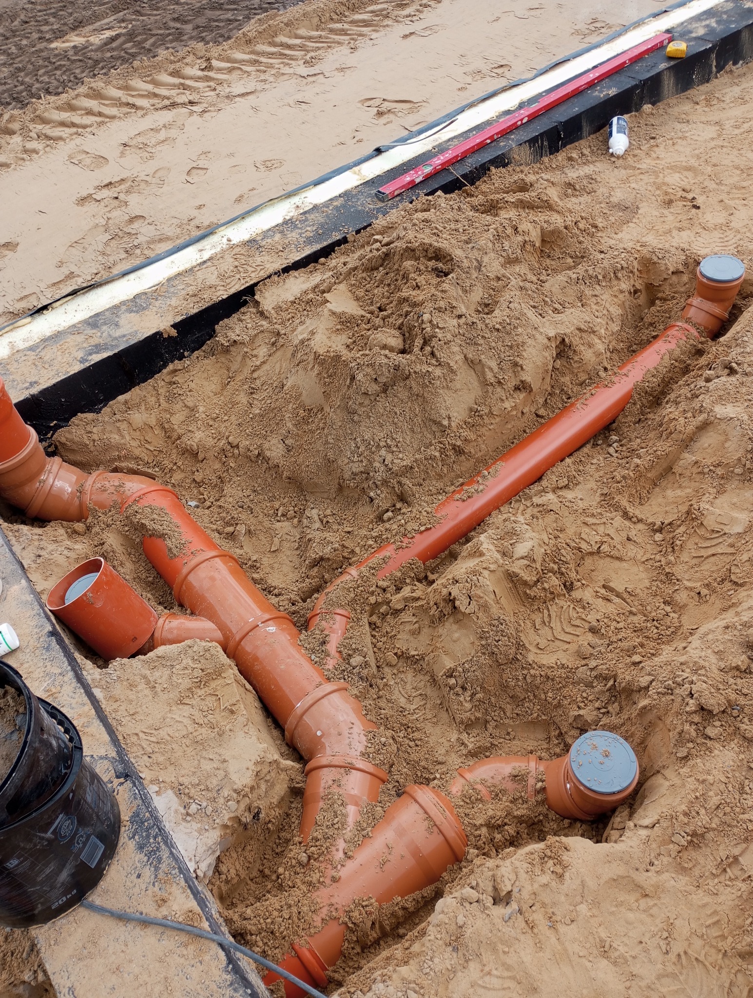 Pomarańczowe rury kanalizacyjne ułożone w wykopie wypełnionym piaskiem, widoczne narzędzia i poziomica na krawędzi wykopu.