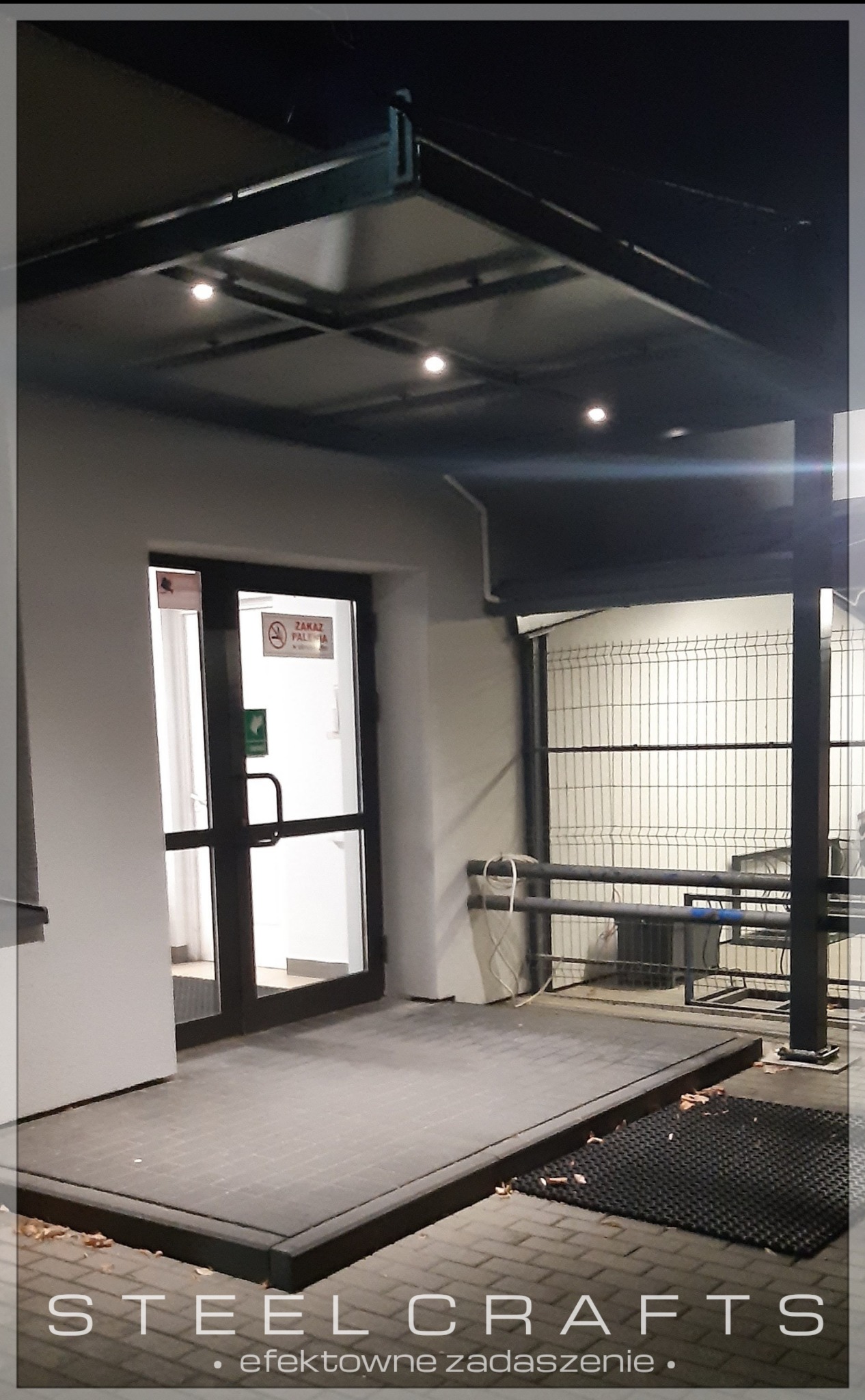 Zadaszenie nad wejściem do budynku z metalową konstrukcją, oświetleniem punktowym i czarnymi ramami drzwi oraz fragmentem ogrodzenia z metalowych prętów w tle.