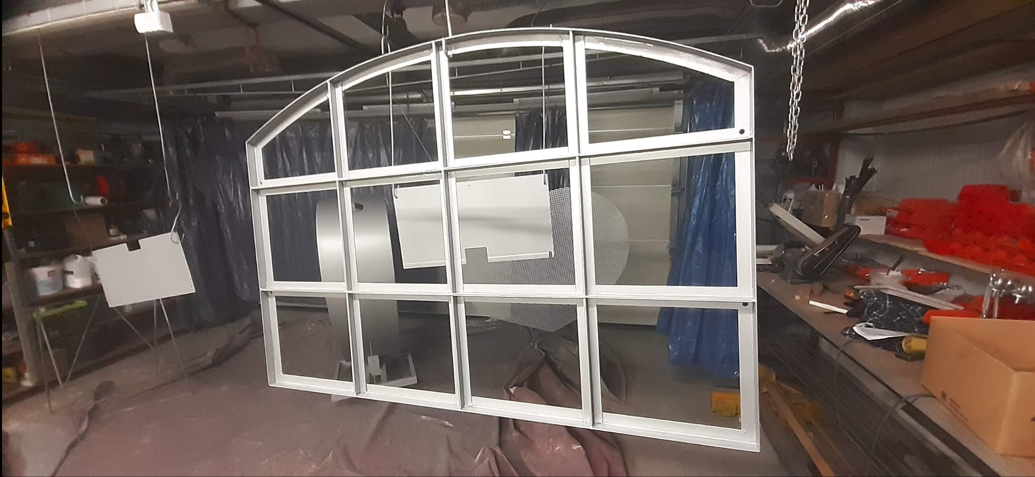 Metalowa konstrukcja z łukiem, pomalowana na biało, zawieszona w warsztacie, widoczne elementy do malowania i narzędzia na stole roboczym.