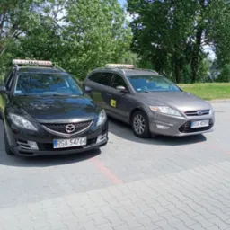Dwa samochody osobowe typu kombi, czarny Mazda i szary Ford, oznakowane jako taksówki z napisami 'LUXTAXI SANOK' na dachu, zaparkowane na parkingu z kostki brukowej, w tle zieleń drzew.