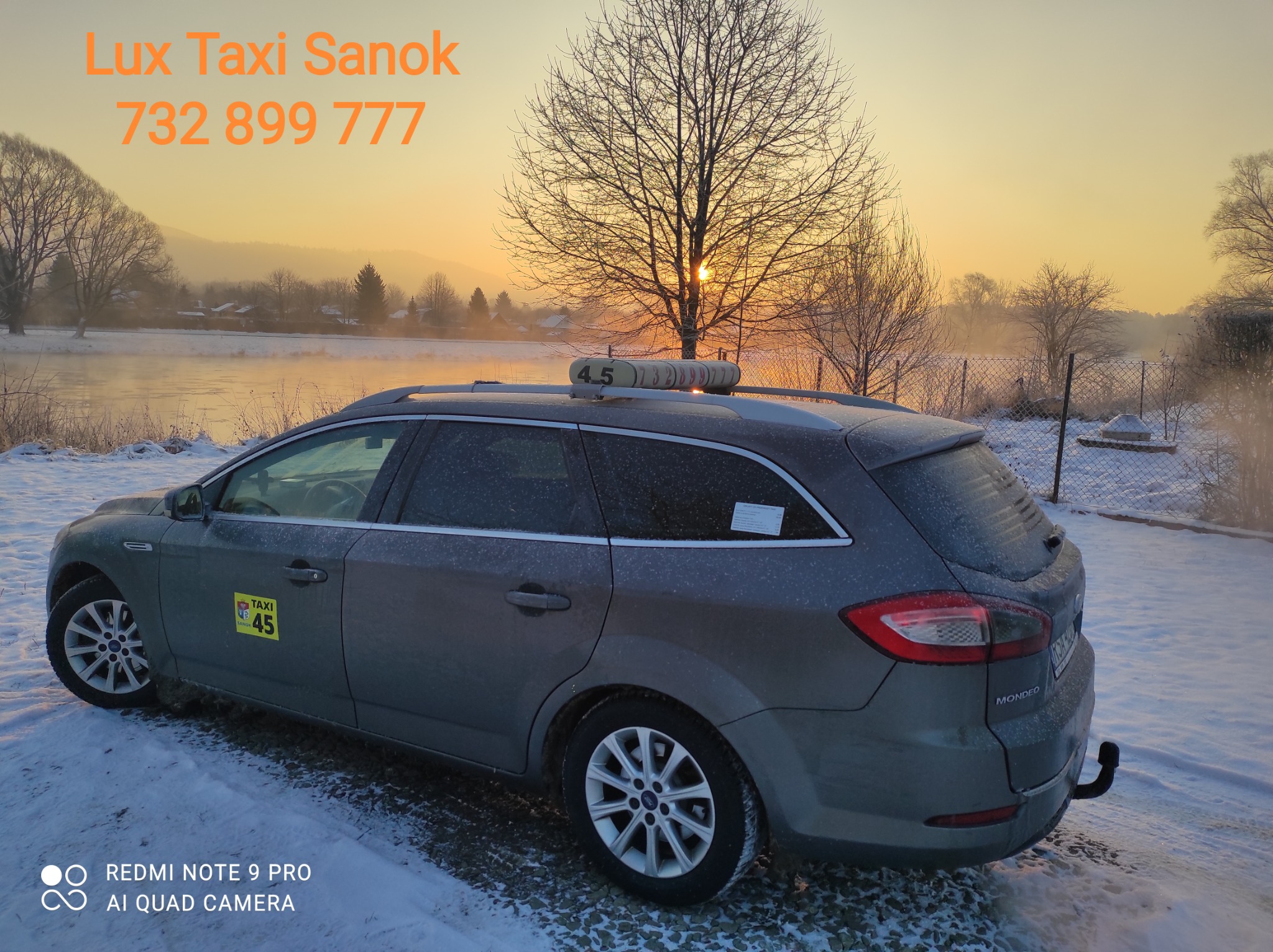 Szary samochód kombi z napisem 'Lux Taxi Sanok' i numerem telefonu, zaparkowany w zimowym krajobrazie o wschodzie słońca