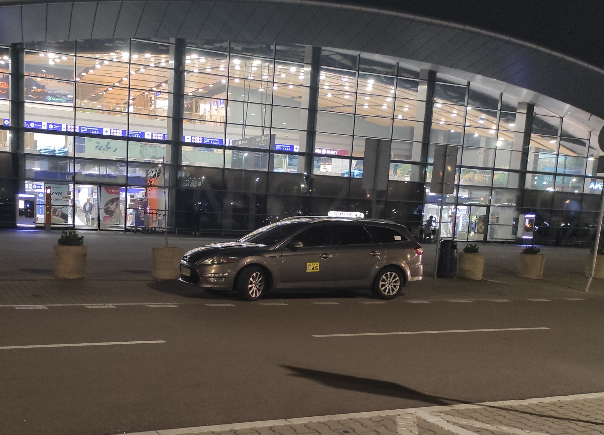 Szary samochód kombi z oznaczeniem Taxi 45 zaparkowany przed przeszklonym budynkiem lotniska nocą.