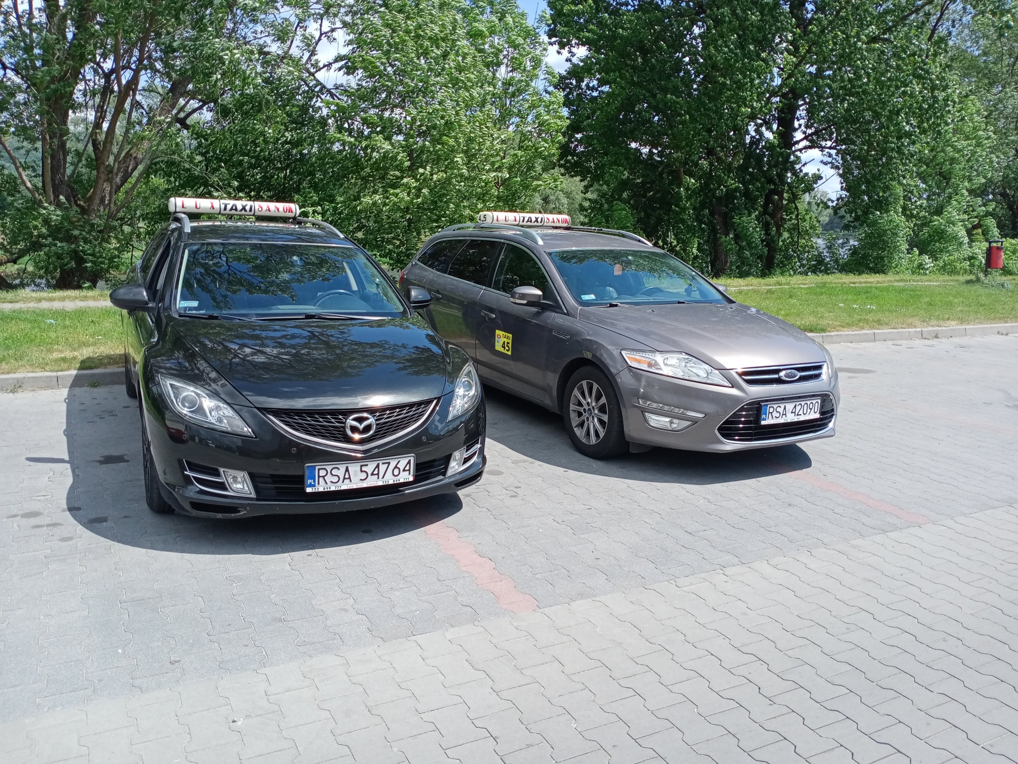 Dwa samochody osobowe typu kombi, czarny Mazda i szary Ford, oznakowane jako taksówki z napisami 'LUXTAXI SANOK' na dachu, zaparkowane na parkingu z kostki brukowej, w tle zieleń drzew.