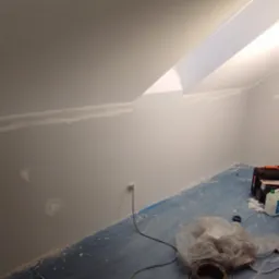 Pok&oacute;j na poddaszu w trakcie szpachlowania, ściany przygotowane pod malowanie, widoczne narzędzia i lampa budowlana.
